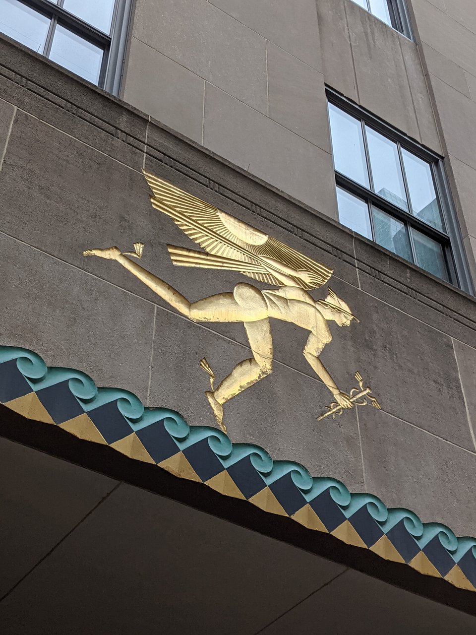 A gold Art Deco relief sculpture depicting a stylized figure of Mercury, messenger of the gods, holding a caduceus, on the façade of a building at Rockefeller Plaza.