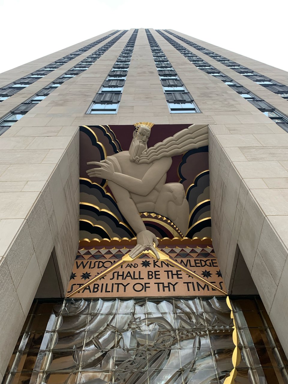 An Art Deco relief sculpture titled ‘Wisdom’ at the entrance of 30 Rockefeller Plaza, depicting a stylized Zeus-like figure rendered with geometric precision and bold lines characteristic of the Art Deco period. The figure holds angular lightning bolts, set against layered, dramatic clouds accented with metallic gold and muted earth tones, accompanied by the inscription ‘Wisdom and Knowledge shall be the stability of thy times’ in a bold, streamlined typeface typical of 1930s design.